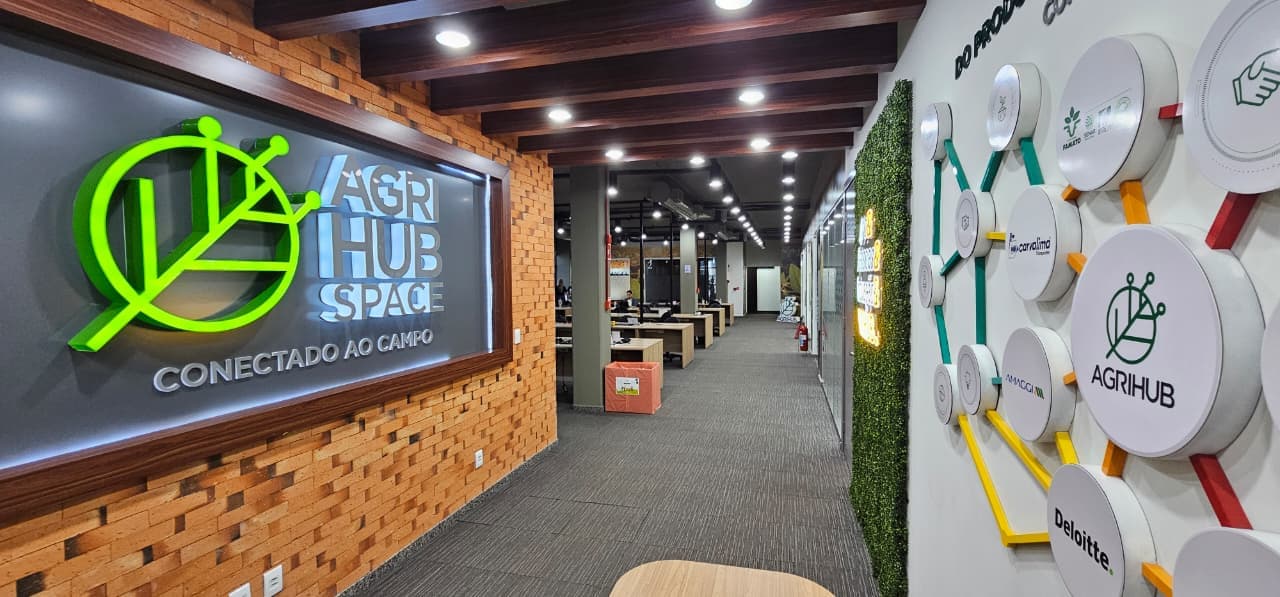 Entrada do AgriHub Space com logo iluminado, parede de tijolos e painel decorativo com logotipos.